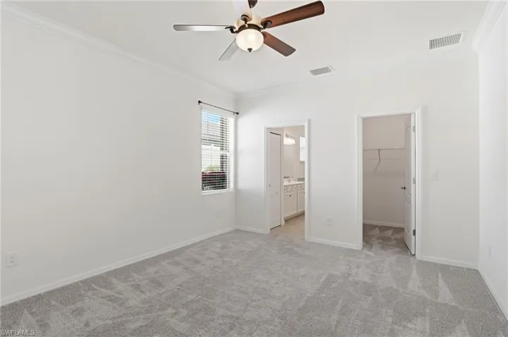 Unfurnished bedroom featuring ornamental molding, a walk in closet, light colored carpet, a ceiling fan, and ensuite bath
