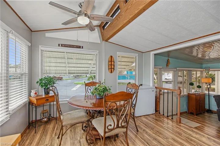 Vaulted ceiling featuring wooden beam accents, wood-finish flooring, ceiling-mounted fan with integrated lighting, and multiple windows with horizontal blinds