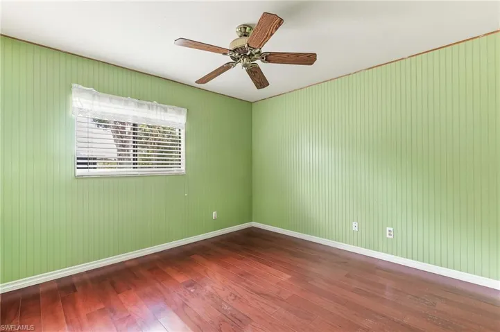 Room featuring rich wood flooring, vertical paneling walls, a window with horizontal blinds and a decorative valance, and an overhead ceiling fan with wood-tone blades