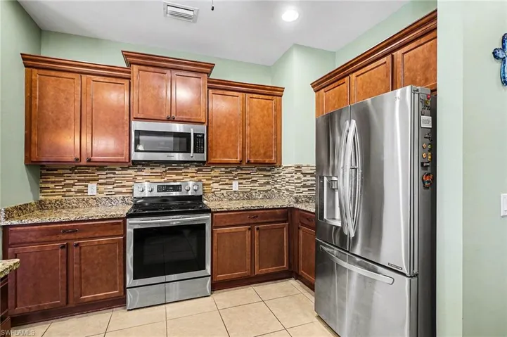 Kitchen with stainless steel appliances, decorative backsplash, light stone counters, brown cabinetry, and light tile patterned flooring