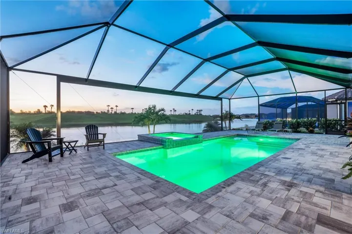 Pool at dusk with glass enclosure, an in ground hot tub, a patio area, and a water view