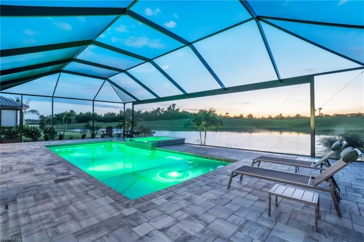 Pool at dusk with glass enclosure, a water view, an in ground hot tub, and a patio area