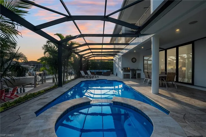 Pool at dusk with area for grilling, a patio area, a pool with connected hot tub, and glass enclosure