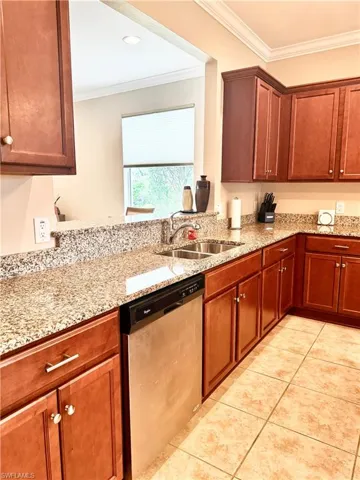 Kitchen featuring crown molding, stainless steel dishwasher, light stone counters, light tile patterned floors, and brown cabinetry