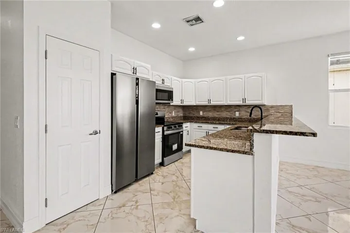 Kitchen with light marble finish floors, stainless steel appliances, a peninsula, white cabinetry, and dark stone counters