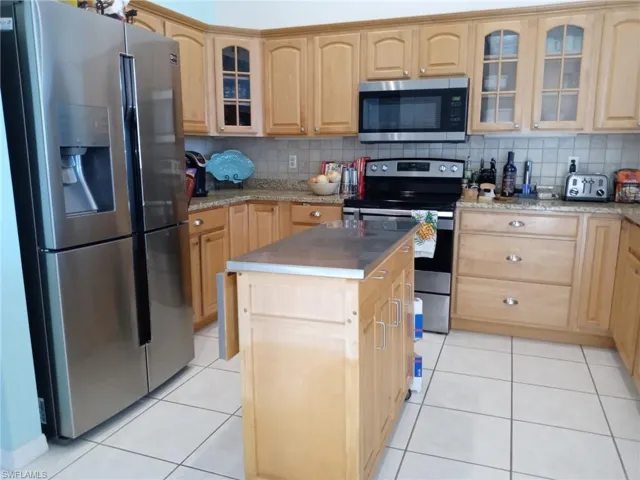 Kitchen with glass insert cabinets, stainless steel appliances, backsplash, light tile patterned floors, and a kitchen island