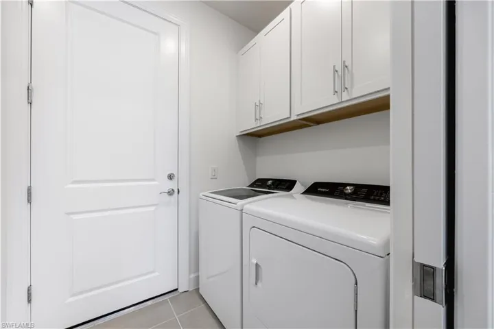 Dedicated laundry area featuring white shaker-style cabinetry with brushed nickel hardware, a wood-finish shelf, and neutral-toned tile flooring