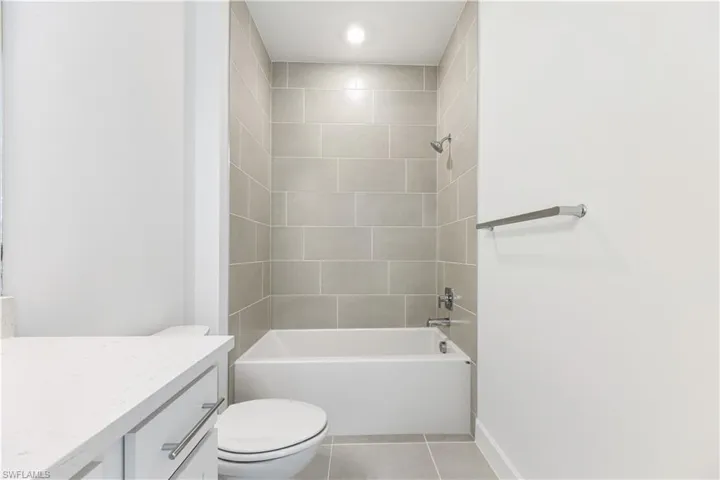 Modern bathroom featuring a built-in tub and shower with light-toned rectangular tile surround, coordinating floor tiles, a white toilet, and a vanity with a white countertop and silver-tone hardware