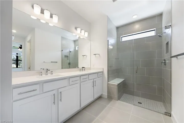 Double vanity with white cabinetry and integrated sinks, a large mirror, and a glass-enclosed shower featuring large format tile, a built-in bench, and a transom window