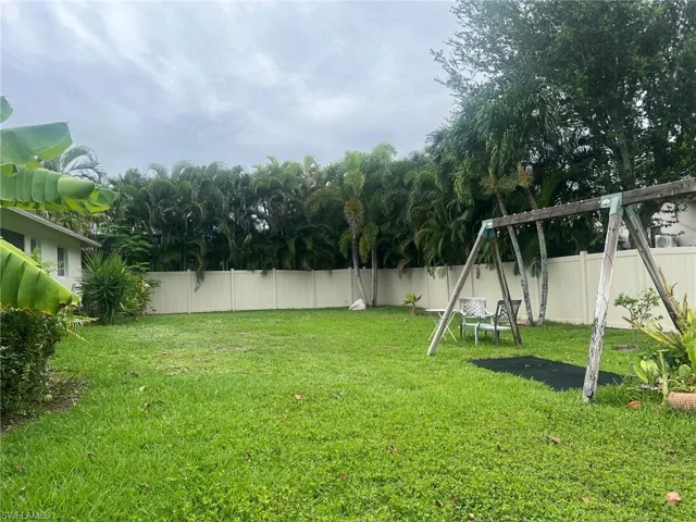 Expansive green lawn area featuring a tall privacy fence, mature tropical foliage, and a wooden playset