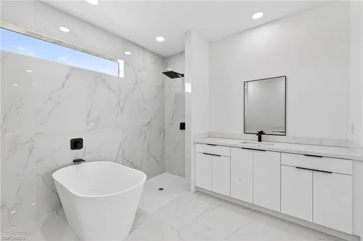 Contemporary bathroom featuring a freestanding soaking tub, a zero-entry shower with a rain showerhead, a dual vanity with white cabinetry and integrated sinks, and white marble-patterned tile flooring and walls