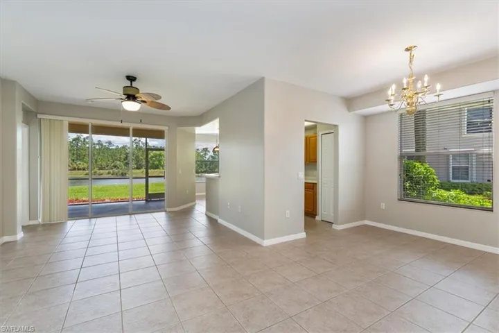 Photo features a bright, open-concept living and dining area with neutral tile flooring and soft beige walls. Large sliding glass doors fill the room with natural light and offer a stunning view of a lush lawn and tranquil waterway. A ceiling fan with leaf-shaped blades adds a touch of Florida charm, while a classic gold chandelier provides elegance over the dining space. The adjoining kitchen is partially visible through a pass-through window and doorway, creating a functional flow between the rooms.