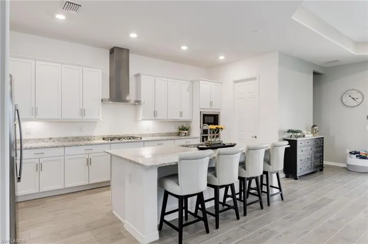 Kitchen with wall chimney exhaust hood, white cabinetry, stainless steel gas cooktop, and an island with sink
