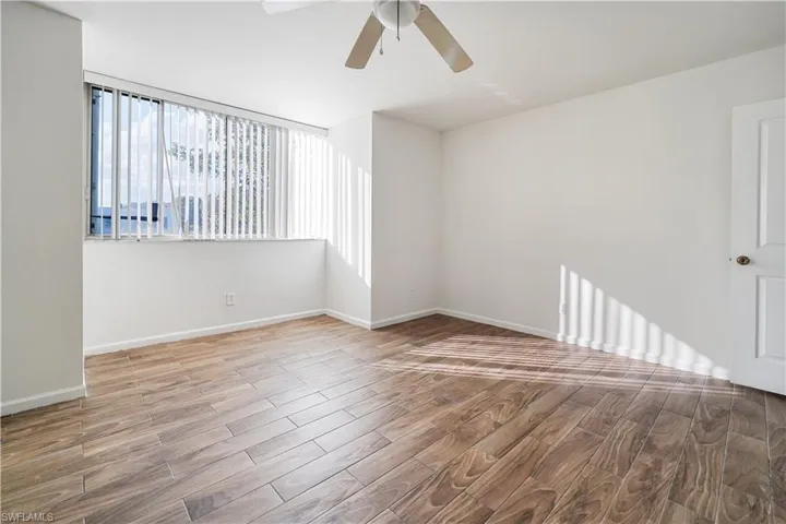 Spacious room featuring wood-finish flooring, a large window with vertical blinds, a ceiling fan, and a white paneled door