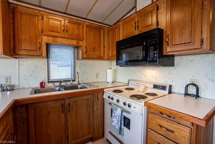 Kitchen with electric stove, brown cabinetry, light countertops, black microwave, and lofted ceiling