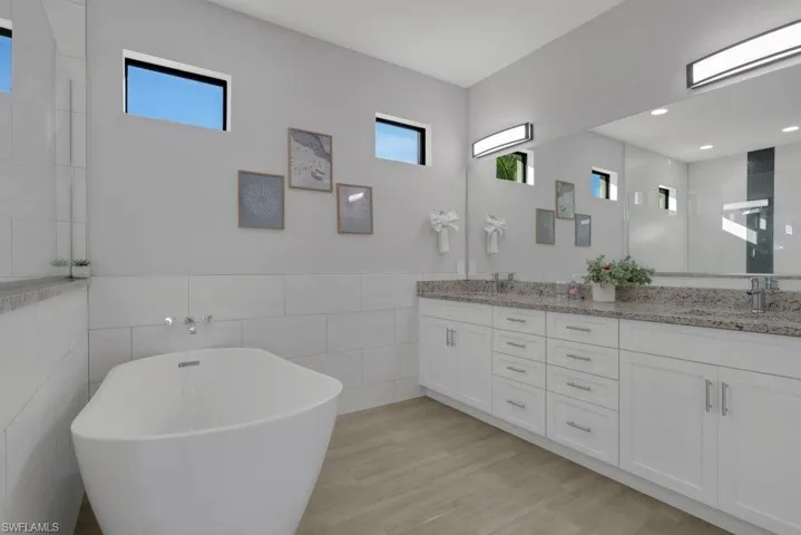 Freestanding soaking tub with wall-mounted faucet, dual vanity with undermount sinks and stone countertop, wood-finish flooring, and horizontal accent windows