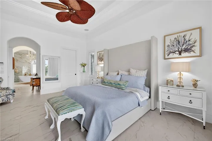 Bedroom featuring light marble finish flooring, arched walkways, a tray ceiling, a ceiling fan, and ornamental molding