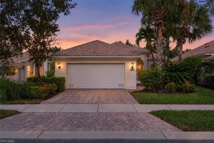 Stucco exterior with a tiled roof, paver driveway, attached two-car garage, and established tropical landscaping
