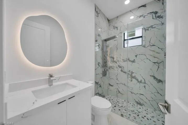 Modern bathroom featuring a custom-shaped backlit mirror, white vanity with integrated sink, and a shower enclosure with floor-to-ceiling marble-look tile