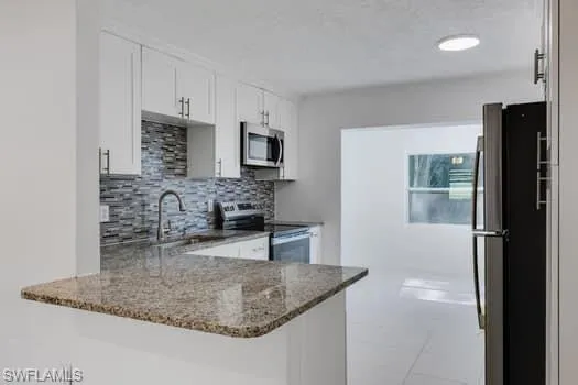 Kitchen with appliances with stainless steel finishes, a peninsula, dark stone counters, backsplash, and white cabinetry