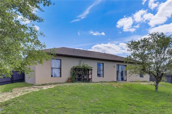 Spacious backyard featuring a stucco exterior, dark shingle roof, multiple windows, and a sliding glass door