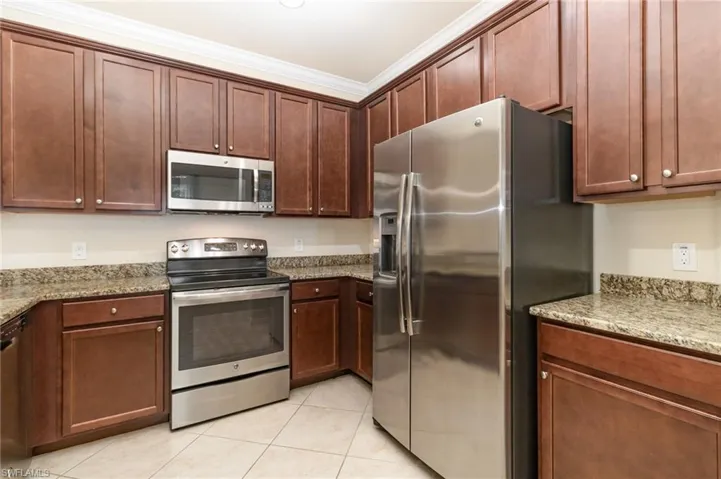 Kitchen with appliances with stainless steel finishes, light stone countertops, ornamental molding, light tile patterned flooring, and dark brown cabinets