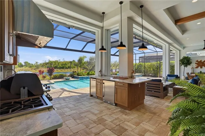 Covered outdoor kitchen featuring stone paver flooring, built-in stainless steel grill, stone-finish countertops, wood-finish cabinetry, and hanging pendant lighting, overlooking a screened-in pool and pond