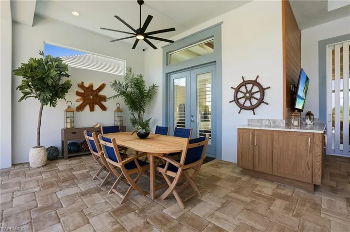 Covered outdoor living space featuring stone paver flooring, a built-in refreshment station with light-tone cabinetry and stone countertop, wood-plank ceiling accent, multi-pane glass doors, and a modern ceiling fan