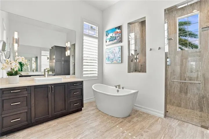 Dark-toned custom cabinetry with integrated surface-mount sink, freestanding soaking tub, stone-finish walk-in shower with glass enclosure, wall-mounted shutters, and large-format wall-to-wall vanity mirror
