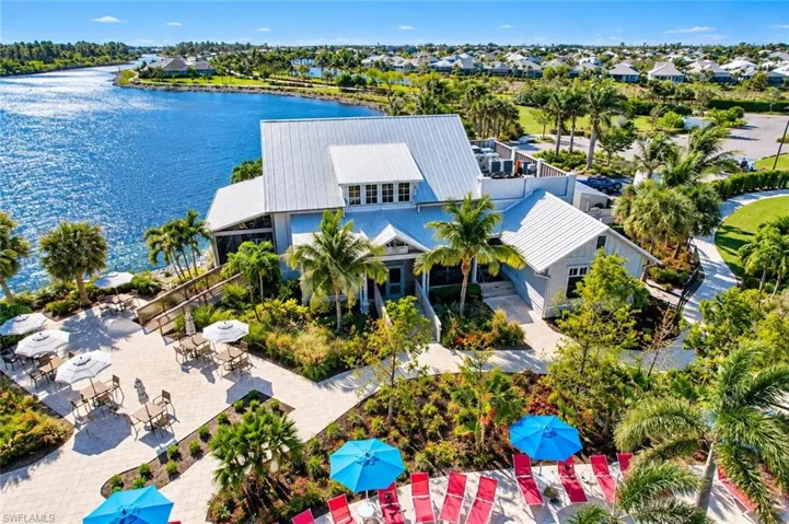 Waterfront clubhouse featuring a metal roof, expansive paver patio, lush tropical landscaping, and mature palm trees
