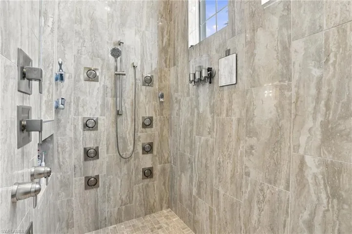 Walk-in shower featuring floor-to-ceiling stone-finish wall tile, multi-head shower system with wall-mounted body sprayers, adjustable handheld shower wand, and inset storage niche