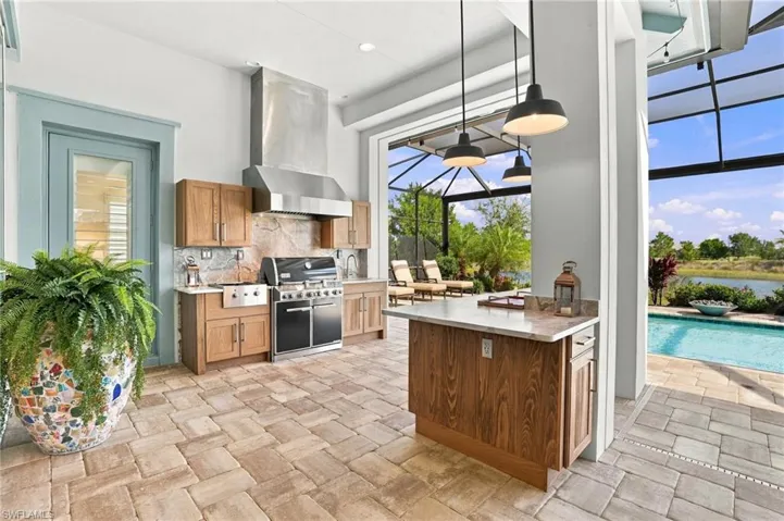 Outdoor kitchen featuring custom wood cabinetry, stainless steel range with overhead vent hood, stone backsplash, stone-paved flooring, and a covered patio area