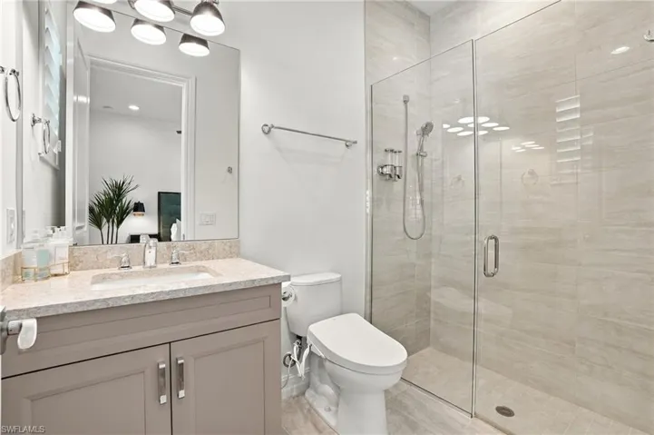 Bathroom featuring a grey vanity with a neutral stone-finish countertop, undermount sink, glass-enclosed walk-in shower with floor-to-ceiling tile, and five-bulb vanity light fixture