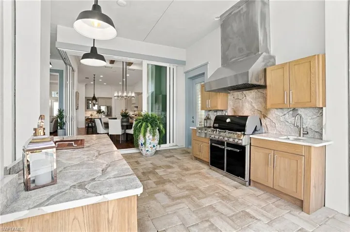 Outdoor kitchen featuring natural stone countertops, custom wood cabinetry, a professional-grade stainless steel range hood, built-in gas grill, and herringbone-patterned stone flooring