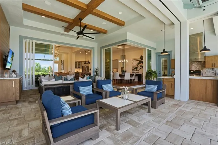Covered outdoor living area featuring a coffered ceiling with wood-finish beams, stone paver flooring, integrated kitchen with wood-finish cabinetry, and large-format retractable glass walls