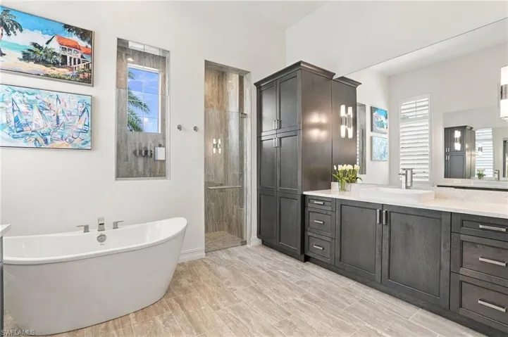 Freestanding soaking tub, walk-in glass-enclosed shower, dark-finish cabinetry with built-in storage, light-toned stone-finish flooring, and expansive wall-mounted vanity mirror