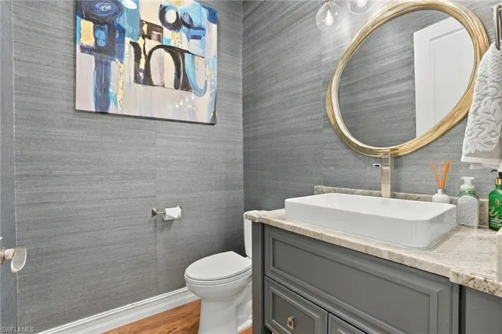 Textured grey wall covering, stone-finish vanity countertop, vessel sink with a tall-profile faucet, oval mirror with a metallic frame, and dark cabinetry