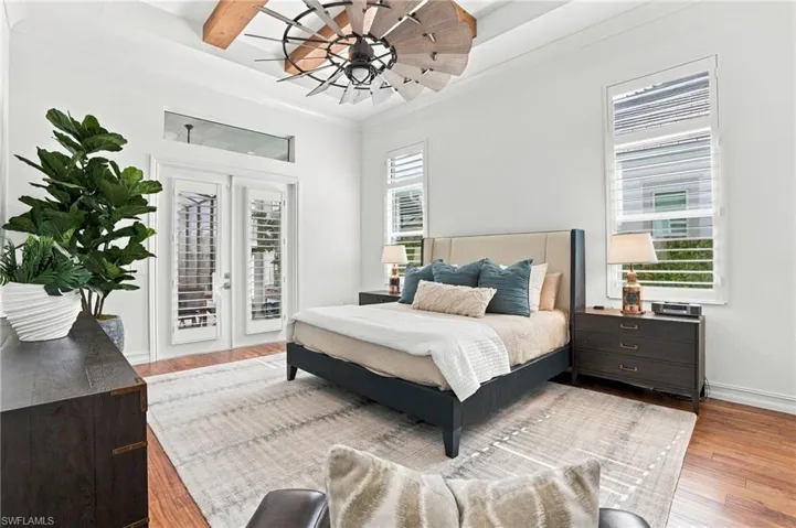 Tray ceiling featuring exposed wood beams, windmill-style ceiling fan, wood-finish flooring, transom window, and plantation shutter window treatments