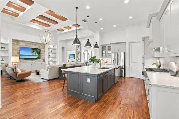 Coffered ceiling with wood-finish beams, stone-accented fireplace wall, expansive center island with countertop seating, shaker-style cabinetry, and wood-finish flooring