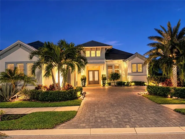 Single-story exterior featuring a paver driveway, double-door entryway, multi-gable roofline, and landscaped grounds with mature palm trees