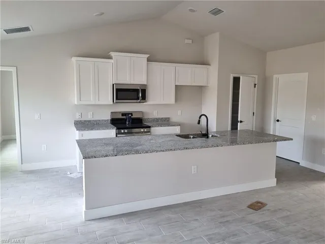 Kitchen featuring stainless steel appliances, light stone counters, white cabinetry, an island with sink, and lofted ceiling