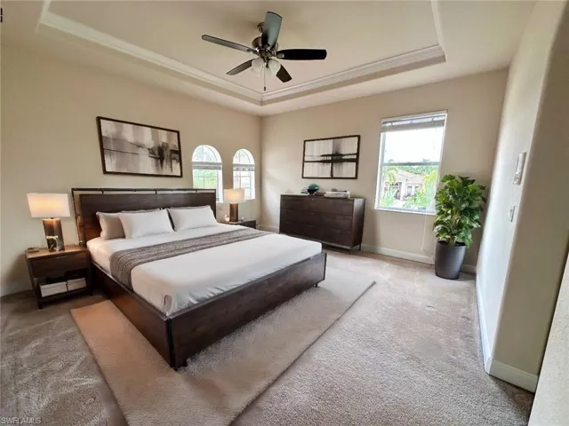 Carpeted bedroom featuring a tray ceiling, multiple windows, ceiling fan, and crown molding