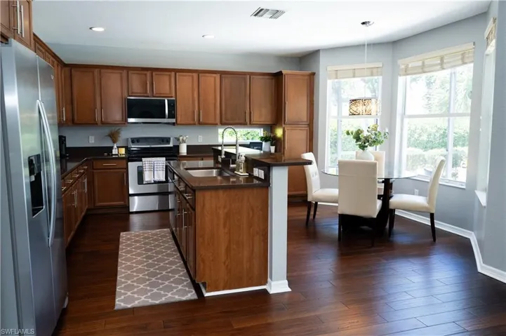 Kitchen featuring wood-finish cabinetry, stainless steel appliances, dark countertops, a central island with an integrated sink, and wood-finish flooring