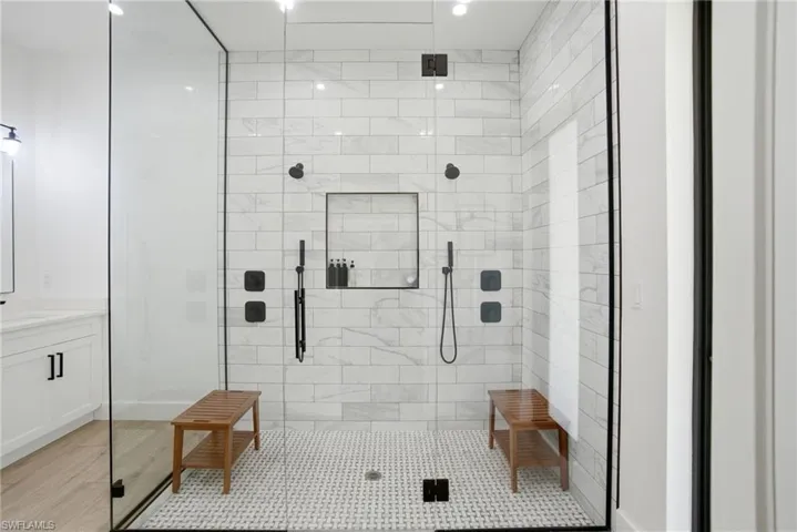 Spacious walk-in shower featuring a glass enclosure with matte black trim, white subway tile walls, a mosaic tile floor, dual shower heads, and an integrated shower niche