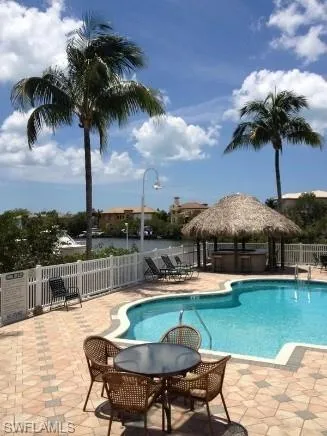 Freeform swimming pool with a surrounding paver patio, featuring a thatched tiki bar and a white picket fence