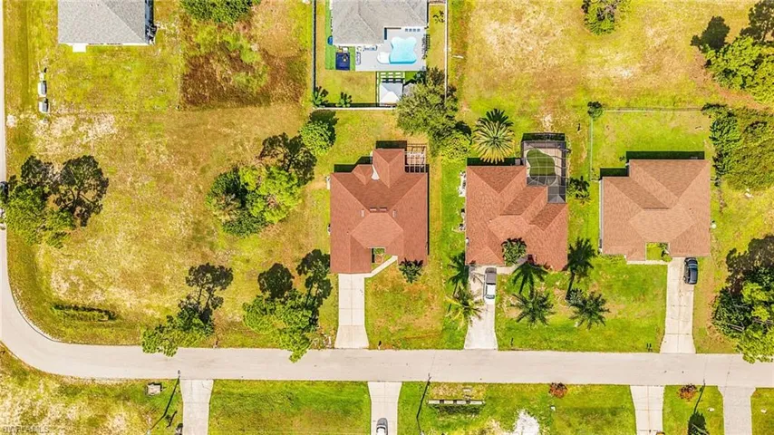 Aerial perspective of a residential street with multiple properties, featuring a corner lot with undeveloped land, houses with brown roofs, and a property with a visible swimming pool