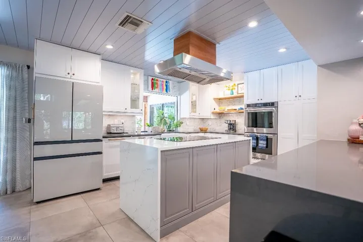 Kitchen featuring a center island with waterfall countertop, stainless steel appliances including a built-in oven and range hood, ample white cabinetry, and wood-finish ceiling panels