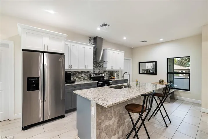 Kitchen featuring an island with sink, white cabinets, wall chimney range hood, appliances with stainless steel finishes, and light stone countertops