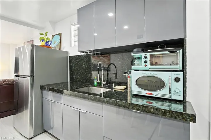 Compact kitchen area featuring a stainless steel refrigerator, dark granite countertops, a stainless steel undermount sink with a matte black faucet, and sleek grey cabinetry