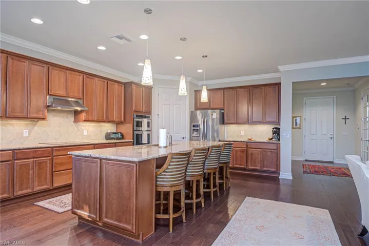 Kitchen with pendant lighting, dark wood-type flooring, appliances with stainless steel finishes, a kitchen island with sink, and light stone counters